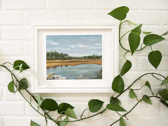 Framed original landscape painting of a blue sky and a hedge of bushes along a rocky river bank.